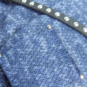 Pode incluir: Close-up de um tecido de brim azul com uma tira de couro preta com tachas prateadas.
