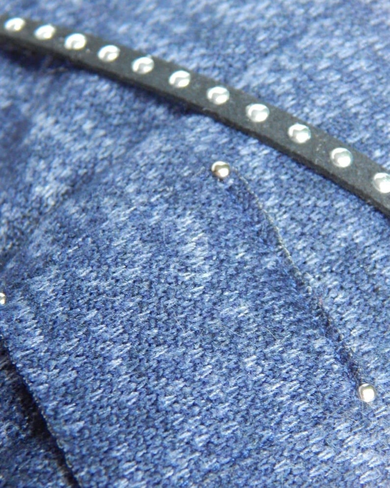 Pode incluir: Close-up de um tecido de brim azul com uma tira de couro preta com tachas prateadas.