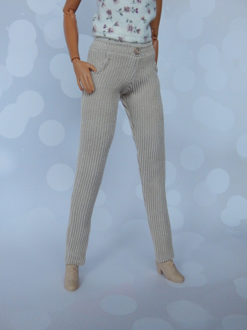 May include: A pair of beige corduroy pants with a button closure and a pocket on the front. The pants are on a doll.