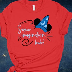 May include: A red t-shirt featuring a whimsical design. The design includes a blue wizard hat with white stars and a crescent moon, black Mickey Mouse ears, and the phrase "Some imagination, huh?" in a playful font.