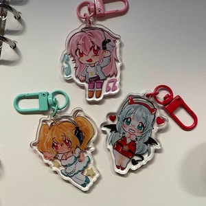 Puede incluir: Tres llaveros acrílicos de estilo anime con diseños coloridos. Un llavero presenta un personaje de pelo rosa con auriculares, otro tiene un personaje con pelo naranja y el tercero tiene un personaje con pelo azul y cuernos de diablo. Cada llavero tiene un cierre de diferente color.