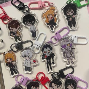 Puede incluir: Una colección de llaveros con personajes de estilo anime. Cada llavero presenta un diseño de personaje diferente, con colores vibrantes e ilustraciones detalladas. Los llaveros están sujetos a cierres metálicos de varios colores.