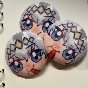 May include: Three round plush buttons featuring a cartoon anime character with blue eyes, gray hair, and a red tongue. The character has a light pink complexion and a yellow and white headpiece. The buttons are on a white surface.