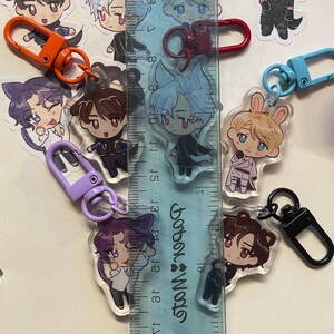 May include: A collection of anime-style acrylic keychains featuring various character designs. Each keychain has a colorful clasp in orange, red, purple, blue, and black. A ruler is placed next to the keychains to measure their size.