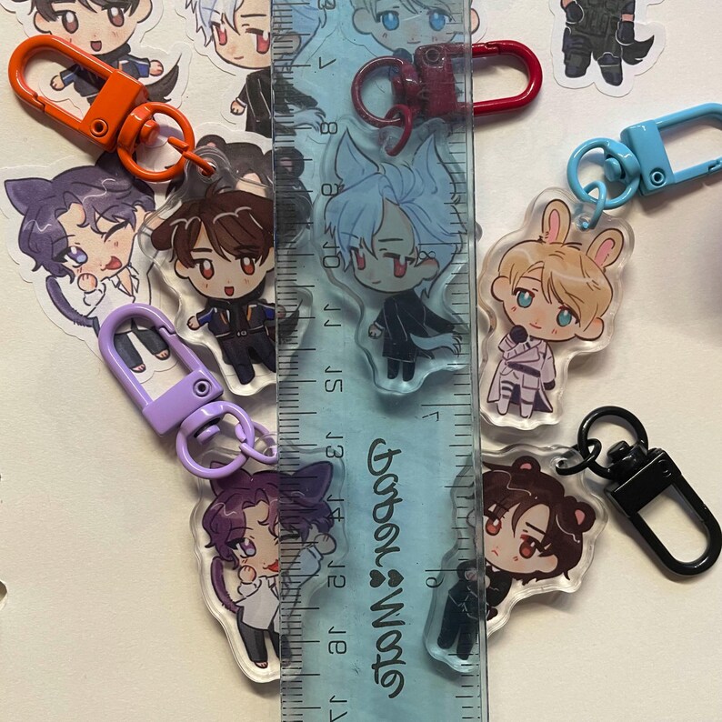May include: A collection of anime-style acrylic keychains featuring various character designs. Each keychain has a colorful clasp in orange, red, purple, blue, and black. A ruler is placed next to the keychains to measure their size.