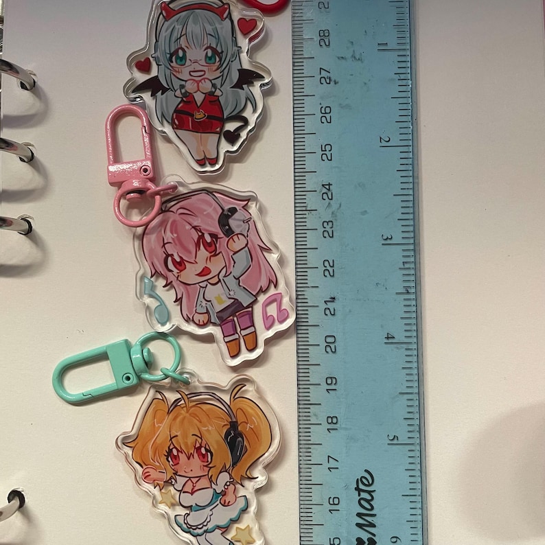 May include: Three anime-style acrylic keychains with colorful illustrations. The keychains feature cartoon characters with headphones, wings, and musical notes. A ruler shows the keychains are approximately 2.5 inches in length.