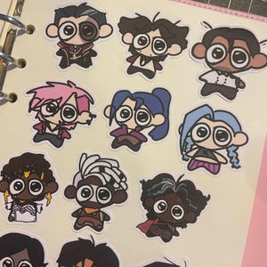May include: A selection of cartoon character stickers with large eyes and varied hairstyles. The stickers display a range of outfits and colours, including pink, blue, and black, against a light background.
