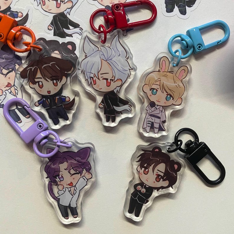 May include: A collection of anime-style acrylic keychains featuring various characters with animal-themed ears and outfits. Each keychain has a different colored clasp, including orange, red, purple, blue, and black. The characters have large eyes and are in various poses.
