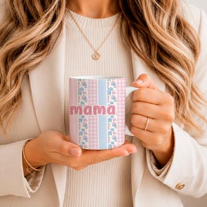 Puede incluir: Una taza de cerámica blanca con un patrón de rayas verticales en azul claro, cuadros vichy rosas y un estampado floral con flores azules y rosas. La palabra mama está impresa en una fuente rosa, audaz y texturizada en el centro del diseño.