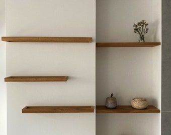 May include: A white wall with a built-in shelf unit. The shelf unit has three wooden shelves and a small niche. The niche contains a small ceramic jar, a woven basket, and a larger woven basket. A small vase with purple flowers sits on the top shelf.
