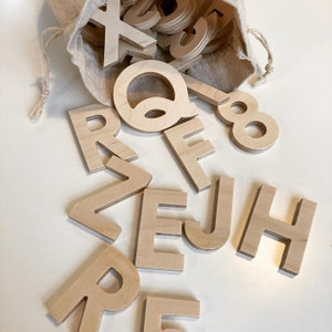 May include: A collection of wooden alphabet letters and numbers, spilling from a beige drawstring bag. The letters are in a sans-serif font and made of light-coloured wood. The letters are scattered on a white surface, with the letter "Q" and the number "8" visible.