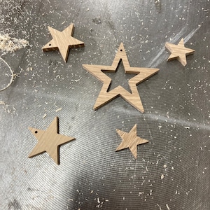 May include: Five wooden star decorations of different sizes, each with a natural wood grain. One star has a cut-out centre. All stars have a small hole for hanging. The stars are scattered on a surface covered in wood shavings.