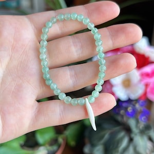 May include: A stretch bracelet composed of small, round, translucent light green beads with subtle internal speckling. The bracelet features a single, smooth, tapered white charm that hangs vertically from the circular beaded strand.