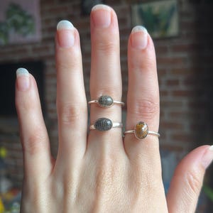 May include: Three silver rings with oval-shaped cabochon stones. Two rings feature dark grey, textured stones with a fine, linear pattern, while the third ring displays a smooth, translucent amber-colored stone. Each ring has a thin, polished band.