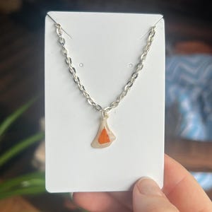 May include: A silver chain necklace with a pendant. The pendant is a light beige shape with an orange triangle in the center. The necklace is displayed on a white card.