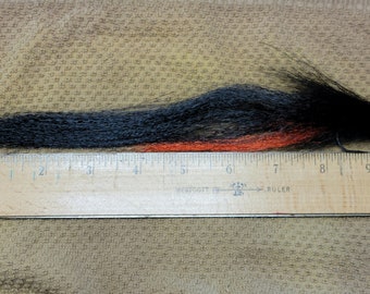May include: A handmade fishing fly with black and orange fibers, measuring approximately 10 inches in length. The fly is positioned next to a wooden ruler, showcasing its size and design. The fly is designed to attract fish.