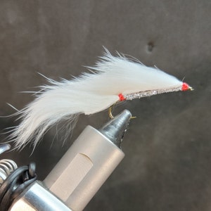 May include: A white fishing fly with a silver body and red accents. The fly is made of soft, fluffy material and has a gold hook. The fly is designed for catching fish and is a popular item for anglers.