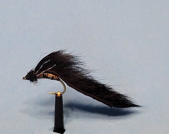 May include: A black and brown fishing fly with a curved hook and a black base. The fly has a long, feathery tail and a small, detailed head. The fly is designed for fishing.