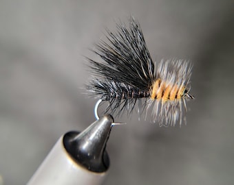 May include: A close-up of a fly fishing lure. The lure features a black and orange body with black and white feather-like extensions. The hook is silver, and the lure is held in a silver clamp against a gray background.