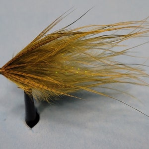 May include: A handmade fishing fly with a golden-brown body and long, flowing feathers. The fly is tied with fine materials and features a small, metallic head. It is mounted on a black stand against a light blue background.