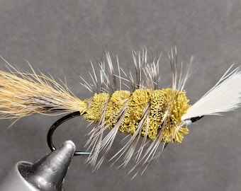 May include: A close-up of a handmade fishing fly. The fly features a black hook with a body of yellow and brown materials, and white feathers. The fly is designed to mimic an insect.