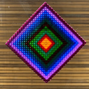 Pode incluir: Um mosaico em forma de diamante colorido feito de pequenas contas de plástico. As contas são dispostas em quadrados concêntricos, com o quadrado central amarelo, rodeado de laranja, vermelho, verde, azul e roxo.