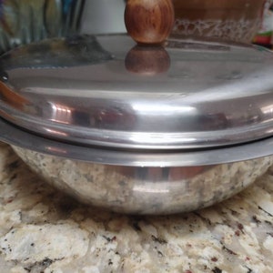 Pode incluir: Uma tigela de servir prateada com tampa e botão de madeira. A tigela tem uma superfície lisa e brilhante e uma forma ligeiramente arredondada.