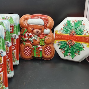 May include: Four red and green Christmas tin boxes with different designs. One box features a Santa's workshop, another has a teddy bear wearing a Santa hat, another has a holly wreath with the text "Seasons Greetings", and the last one has a Christmas tree.
