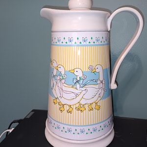 May include: White ceramic pitcher with a lid and handle. The pitcher features a yellow and white striped pattern with a blue border. The design includes illustrations of white geese with blue bows and flowers.