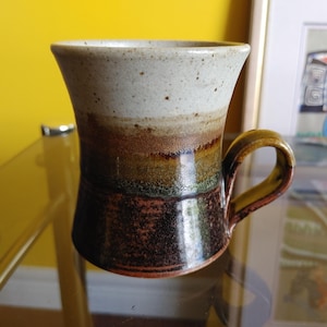 May include: A handmade ceramic mug with a unique, flared shape. The mug features a gradient of colors, including white, brown, green, and dark brown. It has a curved handle and is suitable for hot beverages. The mug is a piece of functional art.