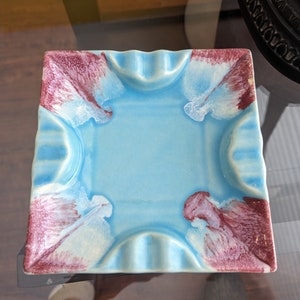 Op de afbeelding: Een vierkante blauwe asbak van keramiek met een gegolfde rand en witte en bordeauxrode marmering.
