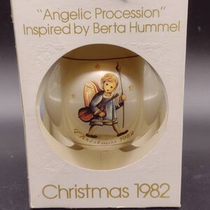 Può includere: Un addobbo natalizio in vetro bianco con un bordo dorato e un'immagine di un angelo che suona uno strumento musicale. L'addobbo è in una scatola con il testo "Angelic Procession" Inspired by Berta Hummel e "Christmas 1982".