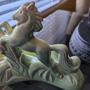 Pu&ograve; includere: Una statuetta in ceramica verde di un cavallo che salta sopra una pianta stilizzata. Il cavallo ha una criniera e una coda dettagliate.