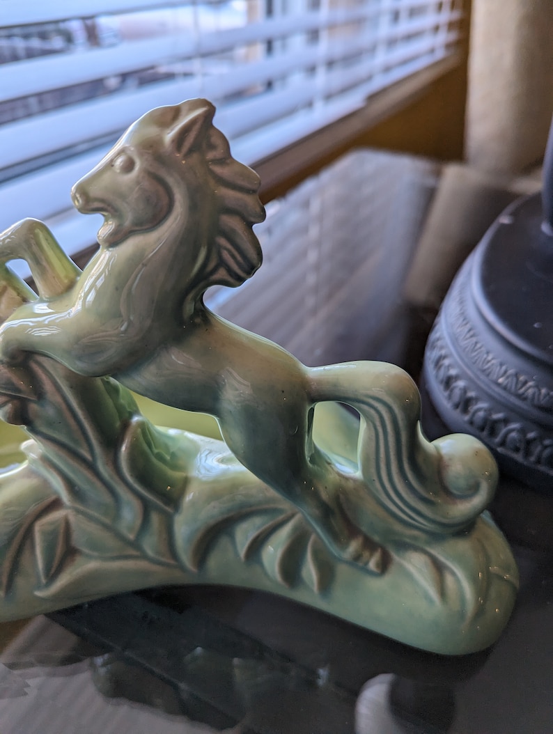 Pu&ograve; includere: Una statuetta in ceramica verde di un cavallo che salta sopra una pianta stilizzata. Il cavallo ha una criniera e una coda dettagliate.