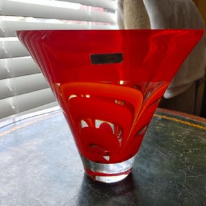 May include: A vibrant red glass vase with a flared, asymmetrical shape. The vase features a clear base and a glossy finish. Swirls of red and white are visible within the glass. A small, rectangular label is affixed to the front of the vase. The vase sits on a dark surface.