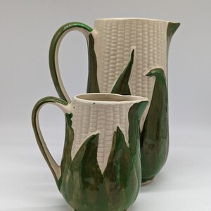 May include: A set of two ceramic jugs with a corn cob design. The larger jug is white with green corn husks and the smaller jug is green with white corn husks.