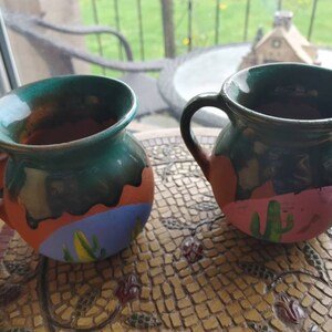 May include: Two green ceramic mugs with painted desert scenes. One mug has a pink desert scene with green cacti, the other has a blue desert scene with yellow cacti.