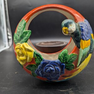 Könnte beinhalten: Ein Keramik-Blumentopf mit rotem Griff und Papageien-Design. Der Topf hat gelbe und blaue Rosen, grüne Blätter und einen braunen Innenraum. Der Papagei ist blau, gelb und grün.