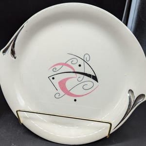 Peut inclure: Deux assiettes en céramique blanche avec une bordure argentée et un motif abstrait noir et rose. Les assiettes sont sur un support en métal doré.