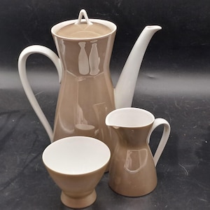 May include: A tan and white ceramic coffee set. The set includes a tall coffee pot with a lid, a creamer, and a small cup. The coffee pot has a white handle and spout, and a design of two bottles. The creamer and cup are tan.