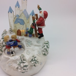 Peut inclure: Une scène de village de Noël en céramique blanche avec un château bleu et blanc, un Père Noël et deux enfants qui patinent sur glace. La scène est éclairée d'une lueur chaude.