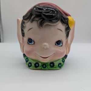 Könnte beinhalten: Eine Keramik-Elfenfigur mit roter Mütze, schwarzen Haaren und blauen Augen. Der Elf trägt einen grün-schwarz gemusterten Kragen.