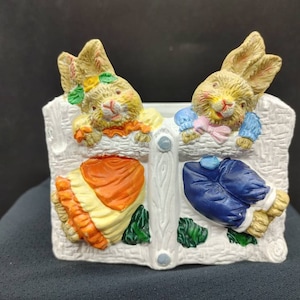 May include: A white ceramic planter featuring two bunny figurines. The bunny on the left is wearing a yellow and orange dress, while the bunny on the right is wearing a blue shirt and trousers. Both bunnies are peering over a white picket fence.