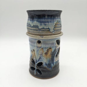 May include: A ceramic candle holder with a blue and brown glaze. The holder has a round top and a cylindrical base with cut-out designs.