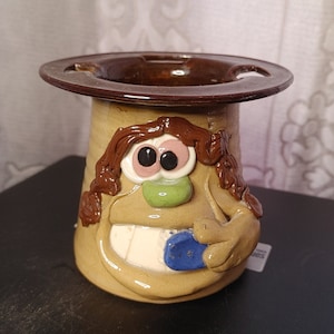 Può includere: Un porta spazzolino da denti in ceramica a forma di viso di cartone animato con capelli castani, occhi grandi e un ampio sorriso. Il porta spazzolino ha un bordo marrone e uno spazzolino da denti blu all'interno.