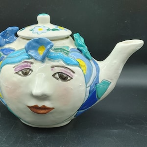 Op de afbeelding: Een witte theepot van keramiek met een gezicht, blauwe en groene bloemdetails en een wit deksel. De theepot heeft een tuit en een handvat.