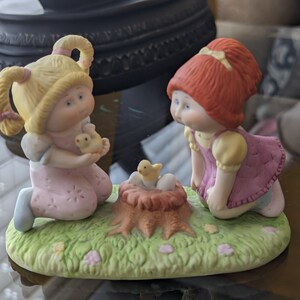May include: A porcelain figurine set featuring two Cabbage Patch Kids dolls. The dolls are kneeling on a green grassy base with a nest of chicks in the center. One doll has blonde hair and is wearing a pink dress. The other doll has red hair and is wearing a pink and yellow dress.