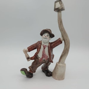 May include: A ceramic figurine of a man holding a bottle and leaning against a lamppost. The man is wearing a brown jacket, a red and white patterned tie, and brown pants. The lamppost is brown and white.