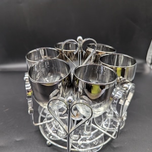 Puede incluir: Un soporte de metal plateado con seis vasos de chupito plateados. El soporte tiene un diseño de corazón y los vasos están dispuestos en círculo.