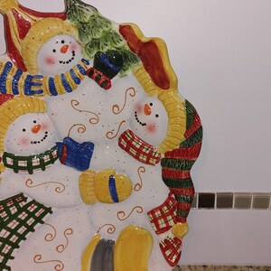 May include: A ceramic snowman serving platter featuring three snowmen wearing colourful scarves and hats. The platter is white with a speckled finish and has a green tree design in the background.
